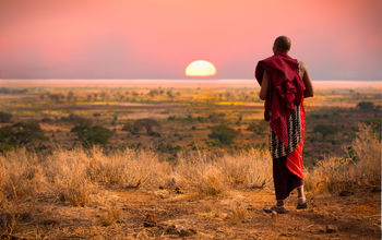 Masai man, wearing traditional blankets, overlooks Serengeti in Tanzania as the colorful sunset fills the sky. Wild grass in the forground. (photo via jocrebbin/iStock/Getty Inages Plus)