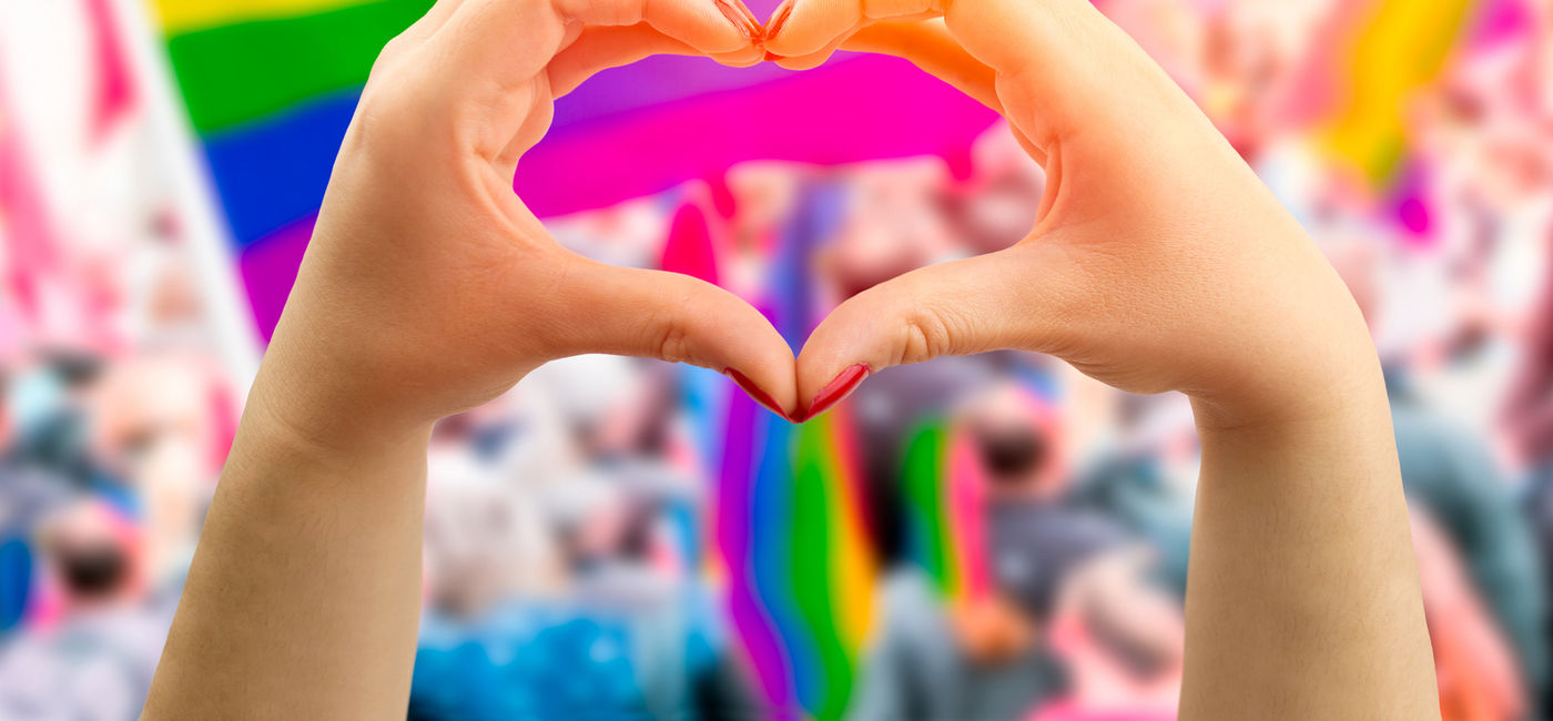 Image: Hands forming the shape of a heart in front of a rainbow flag flying at an LGBTQ+ Pride parade. (photo via iStock/Getty Images Plus/Cunaplus_M.Faba)