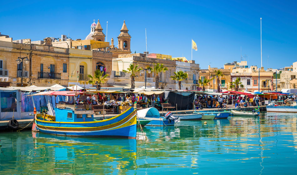 Malta - Marsaxlokk market with traditional Luzzu fishing boats on a beautiful summer day withblue sky and green sea (photo via ZoltanGabor / iStock / Getty Images Plus)