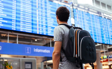 Young man with backpack in airport near flight timetable (Photo via furtaev / iStock / Getty Images Plus)